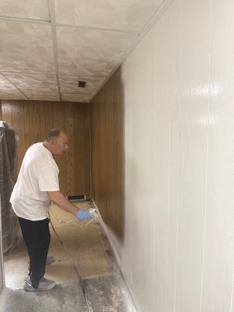 A man in a white shirt and gloves sprays a fresh coat of paint on a wooden wall