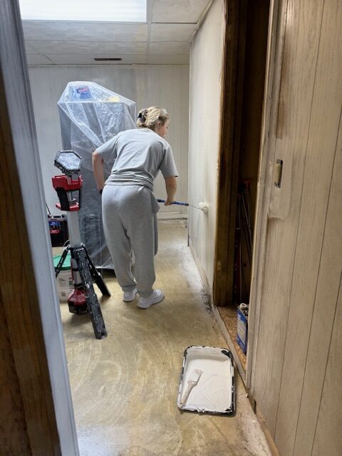 A person in gray sweats paints a wall with a roller in a hallway, surrounded by painting tools and covered furniture, indicating a home renovation project.