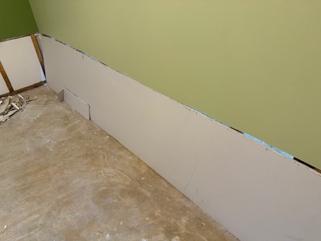 Drywall installation nearing completion, with a green-painted wall and debris on the floor, showcasing progress in a renovation project.