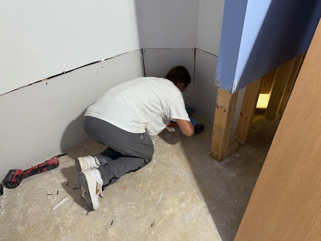 Person kneeling in a room, using a power tool to install drywall while surrounded by construction materials. Renovation in progress.