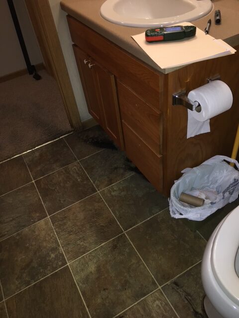 Bathroom floor showing water damage near a wooden cabinet, with a toilet and trash can visible. Relevant for discussing home maintenance issues.